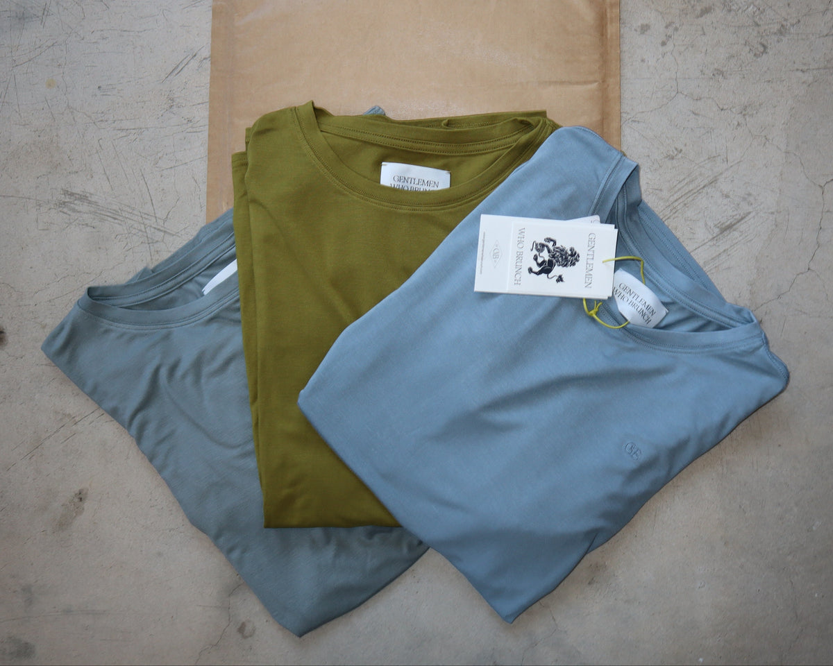 Three folded crewneck t-shirts in muted blue, olive green, and gray are stacked on top of a brown paper mailer bag, laid flat on a textured concrete surface. Each shirt has a hangtag attached.